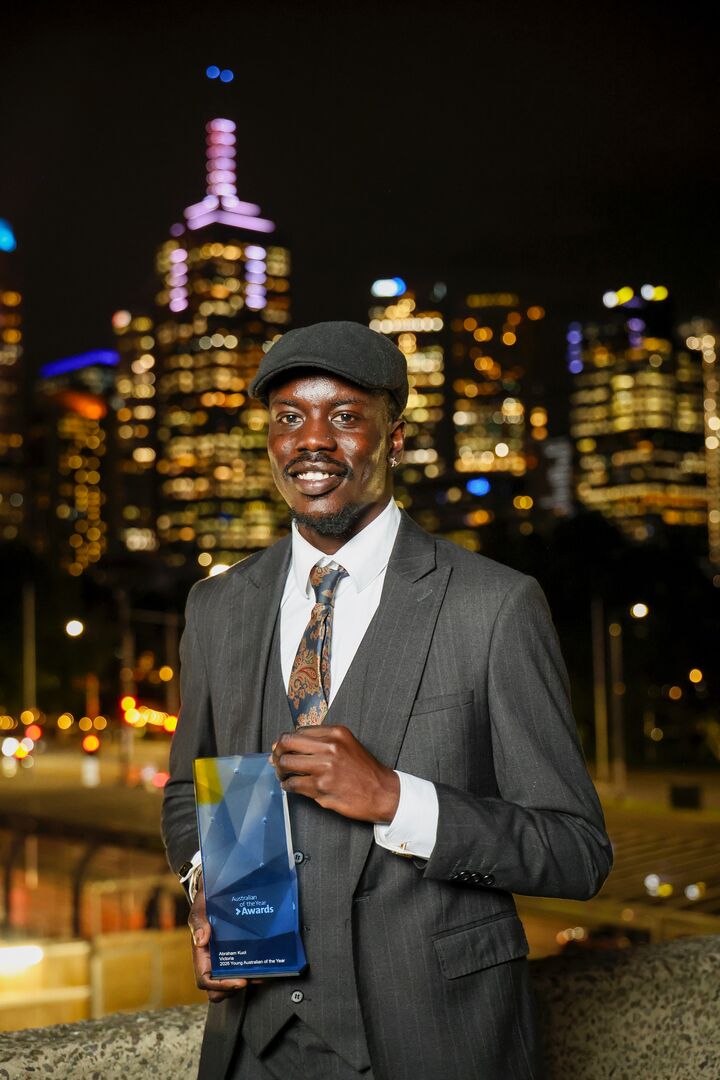 Abraham Kuol is photographed at night, holding his Victorian Young Australian of the Year Award with a city nightscape in the background